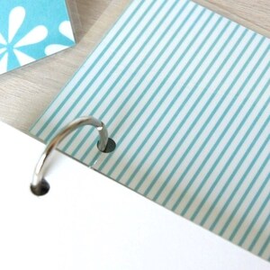May include: Close-up of a notebook with a silver binder ring. The pages have a white background with light blue stripes and a floral pattern. The notebook is on a light wood surface.