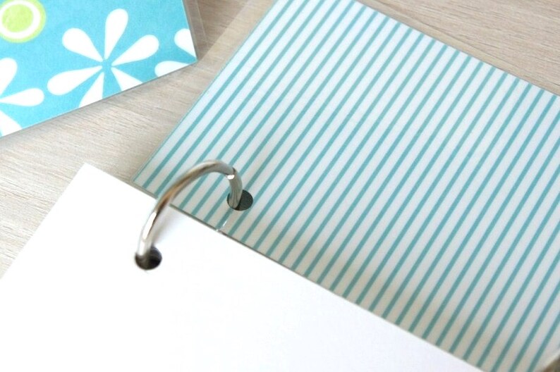 May include: Close-up of a notebook with a silver binder ring. The pages have a white background with light blue stripes and a floral pattern. The notebook is on a light wood surface.