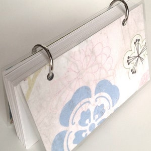 May include: A small notebook with floral patterned pages, held together by two silver metal binder rings. The floral pattern features light blue, pink, and green designs on a white background. The notebook is encased in a clear plastic cover.