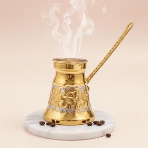 Könnte beinhalten: Eine goldfarbene türkische Kaffeekanne mit einem verzierten Blumendesign und einem langen Griff. Dampf steigt aus der Kanne auf, was auf frisch gebrühten Kaffee hindeutet. Die Kanne steht auf einem weißen Marmoruntersetzer, mit Kaffeebohnen darum verstreut.