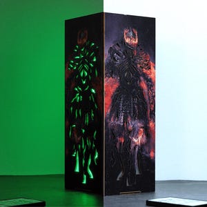 May include: A wooden lamp with a black and red design of a figure with a crown. The lamp is lit up with a green light on the left side.