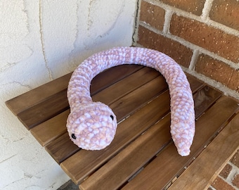 May include: A handmade, plush snake toy in shades of pink and white, with small black eyes. The snake is curled on a wooden folding table. The soft, textured material gives the snake a cuddly appearance.