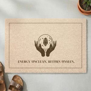 Op de afbeelding: Een rechthoekige beige deurmat met een donkerbruine afbeelding van twee handen die een kristallen bol met sterren vasthouden. De tekst "ENERGY UNCLEAN, RETURN UNSEEN." staat in donkerbruin onder de afbeelding, omringd door een dunne donkerbruine rand.