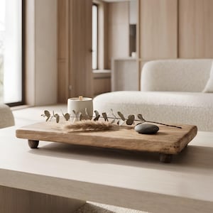 以下が含まれることがあります： 4つの小さな丸い脚が付いた長方形の木製ライザートレイ。トレイの上には、白い円筒形のキャンドル、緑の葉が付いた乾燥したユーカリの枝、ベージュのパンパスグラスの房、滑らかな濃い灰色の川石が置かれています。