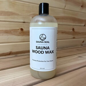 May include: A clear plastic bottle with a black cap, filled with a light yellow liquid. The label reads "SAUNA SEAL SAUNA WOOD WAX" with a logo of a bird. The bottle contains 32 fl oz of product. The background is wooden planks.