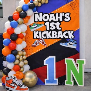 May include: A colorful party backdrop with the text "Noah's 1st Kickback" and sneaker illustrations. A balloon arch in orange, black, white, gold, and blue frames the scene. Large cutouts spell out "1N" in blue and green.