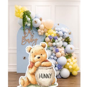 May include: A Winnie the Pooh-themed baby shower decoration. The scene features a cutout of Winnie the Pooh with a honey pot, a blue arch with the words "Oh Baby", and a balloon arch in pastel colors with bee accents.