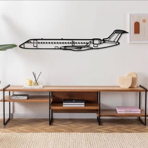 May include: Black metal airplane wall decor with a minimalist design. The airplane is mounted on a white wall above a wooden shelf with books and decorative objects.