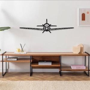 May include: A black metal airplane wall art piece. The airplane is mounted on a white wall above a wooden console table with black metal legs. The room is decorated with a plant, candles, and artwork.