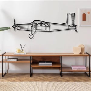 May include: Black metal wall art depicting a vintage airplane silhouette. The airplane is mounted on a white wall above a wooden shelf with black metal legs. The shelf is decorated with books, candles, and decorative objects.