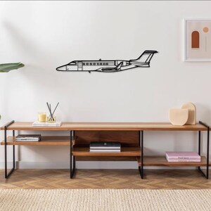 May include: A black metal wall art piece depicting a detailed airplane silhouette. The artwork is mounted on a white wall above a wooden console table with black metal legs. The room is decorated with a large plant, candles, and decorative objects.