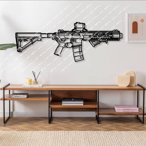May include: Black metal wall art depicting a rifle with a magazine and a scope. The art is mounted on a white wall above a wooden shelf with black metal legs. The shelf is decorated with books, decorative objects, and a pink book.
