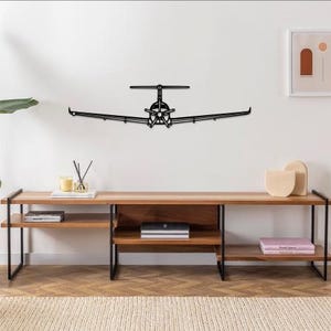 Könnte beinhalten: Ein Wandkunstwerk aus schwarzem Metall, das ein Flugzeug darstellt. Das Kunstwerk ist an einer weißen Wand über einem Medienmöbel aus Holz angebracht. Das Flugzeug hat ein detailliertes Design, einschließlich Flügeln, Rumpf und Propeller.