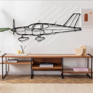 May include: Black metal wall art depicting a small airplane. The artwork is mounted on a white wall above a wooden console table with black metal legs. A framed abstract print and decorative items are also on the wall and table.