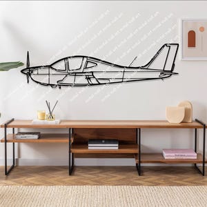 Op de afbeelding: Zwarte metalen vliegtuig wandkunst. Het kunstwerk toont het zijaanzicht van een klein vliegtuig met een propeller. Het vliegtuig is gemonteerd op een witte muur boven een houten console met zwarte metalen poten. De kamer is ingericht met planten en minimalistische decoratie.