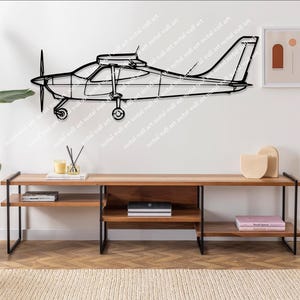 Op de afbeelding: Zwarte metalen wandkunst die een klein vliegtuig afbeeldt. De omtrek van het vliegtuig omvat de propeller, vleugels en staart. Het kunstwerk is bevestigd aan een witte muur boven een houten console, ongeveer 180 cm lang.