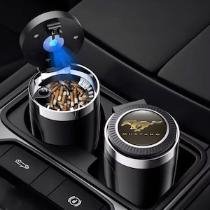 Puede incluir: Cenicero de coche negro con borde plateado y luz azul. El cenicero tiene tapa y está abierto, revelando colillas de cigarrillos. También se ve un segundo cenicero con el logotipo de Mustang.