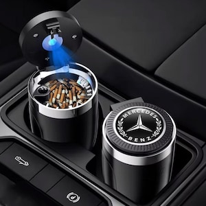 以下が含まれることがあります： シルバーのトリムと青いライトが付いた黒い車の灰皿。灰皿は開いており、タバコの吸い殻が見えます。メルセデス・ベンツのロゴが入った2つ目の灰皿が隣にあります。灰皿は車のコンソールにあります。