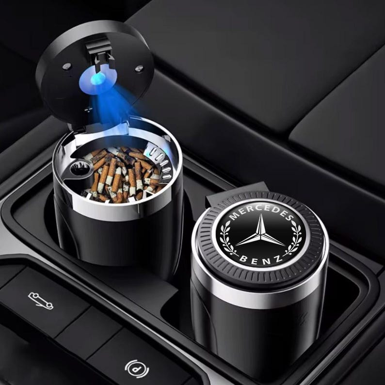以下が含まれることがあります： シルバーのトリムと青いライトが付いた黒い車の灰皿。灰皿は開いており、タバコの吸い殻が見えます。メルセデス・ベンツのロゴが入った2つ目の灰皿が隣にあります。灰皿は車のコンソールにあります。