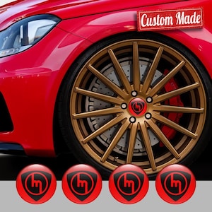 Puede incluir: Primer plano de una rueda de coche roja con llanta dorada y neumático negro. La rueda tiene un tapacubos personalizado con un logotipo negro sobre fondo rojo. El texto "Custom Made" es visible.