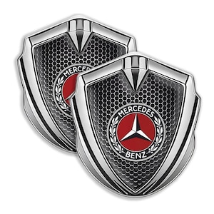 Könnte beinhalten: Zwei verchromte Mercedes-Benz-Emblem-Abzeichen. Jedes Abzeichen hat eine schildförmige Form mit einem schwarzen Wabenmuster, einem roten Kreis mit einem weißen Stern und den Worten "MERCEDES BENZ".