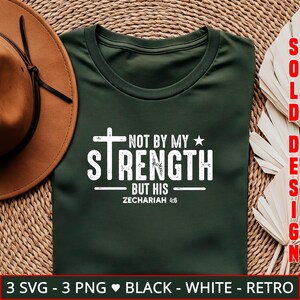 K&ouml;nnte beinhalten: Dunkelgr&uuml;nes T-Shirt mit dem wei&szlig;en Text "NOT BY MY STRENGTH BUT HIS" mit einem Kreuz- und Sterndesign. Der Text "ZECHARIAH 4:6" ist ebenfalls enthalten. Auf der linken Seite des Shirts befindet sich ein brauner Hut. Die Worte "SOLD DESIGN" stehen auf der rechten Seite.