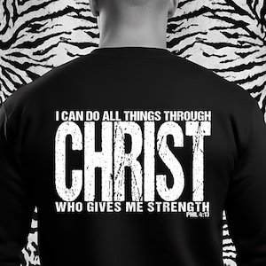 Puede incluir: Una sudadera negra de manga larga con texto blanco de estilo desgastado en la parte posterior que dice I CAN DO ALL THINGS THROUGH CHRIST WHO GIVES ME STRENGTH PHIL 4:13. La prenda tiene un cuello redondo y una construcción de tela negra.