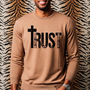 K&ouml;nnte beinhalten: Ein beigefarbenes Sweatshirt mit Rundhalsausschnitt und dem Wort "TRUST" in schwarzen Blockbuchstaben. Das Wort "TRUST" wird von den S&auml;tzen "IN THE LORD", "WITH ALL" und "YOUR HEART" in vertikaler Anordnung begleitet. Der Hintergrund ist ein Tigerprint.