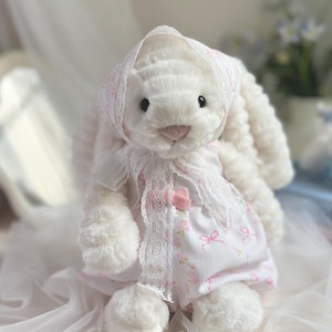 Puede incluir: Un conejo de peluche blanco con un vestido rosa con estampado floral y un gorro a juego. El vestido está adornado con encaje blanco y un lazo rosa. El conejo tiene una nariz rosa y ojos negros, creando una estética suave y encantadora.
