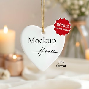 Puede incluir: Un adorno blanco en forma de corazón con el texto "Mockup House" en escritura negra, suspendido por una cuerda dorada. Una pegatina roja "BONUS Feature Image" está adjunta. El fondo incluye velas y flores.