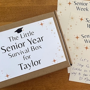 May include: A graduation-themed gift box labeled "The Little Senior Year Survival Box for Taylor" with accompanying cards. Cards include "Happy Graduation Day! 2026" and "Senior Year Week" notes. A five-dollar bill is also included.