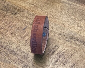 May include: Brown leather bracelet with the text "#Asheville" engraved on it.