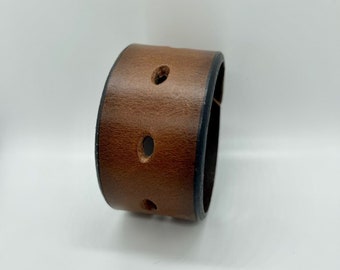 May include: A brown leather cuff bracelet with three evenly spaced holes. The bracelet has a dark edge and a slightly distressed appearance. The cuff is a wide band, designed to be worn around the wrist.