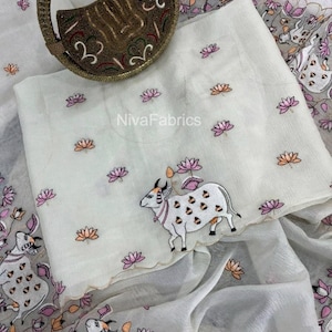 Könnte beinhalten: Ein weißer Stoff mit gestickten rosa und orangefarbenen Lotusblumen und einer weißen Kuh mit braunen Flecken. Eine runde, dunkelbraune, perlenbesetzte Handtasche mit goldfarbenen Metallakzenten liegt darauf. Der Text NivaFabrics ist mittig sichtbar.