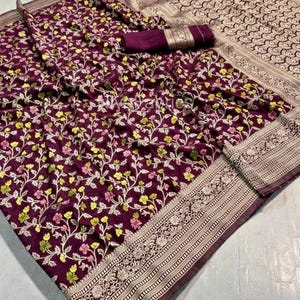 May include: A deep purple saree with a floral pattern in yellow, pink, and white. The border features intricate gold and cream-colored designs. A matching fabric piece is included.