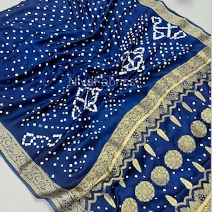 Pode incluir: Um tecido azul marinho com um padrão de pontos brancos e uma borda dourada decorativa. O tecido é drapeado, mostrando o design e a textura intrincados. O texto "Niva fabrics" é visível.