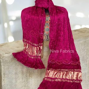 Op de afbeelding: Een magenta sjaal met gouden details en een gefranjerde zoom is over een mannequin gedrapeerd. De sjaal heeft een getextureerde afwerking en is versierd met kleine, witte stippen. De tekst "Niva Fabrics" is zichtbaar.