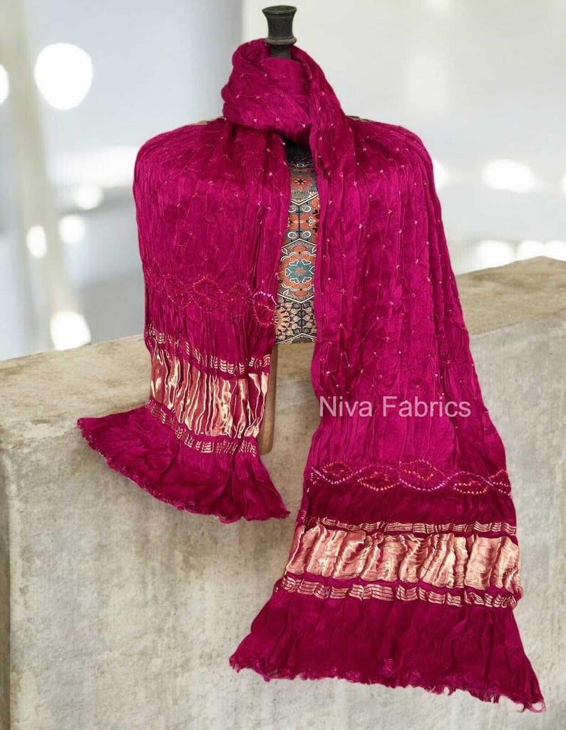 Op de afbeelding: Een magenta sjaal met gouden details en een gefranjerde zoom is over een mannequin gedrapeerd. De sjaal heeft een getextureerde afwerking en is versierd met kleine, witte stippen. De tekst "Niva Fabrics" is zichtbaar.