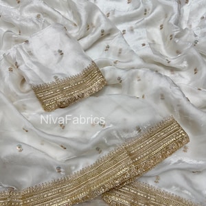 Puede incluir: Tela de color marfil con ribete dorado y delicado bordado floral. La tela está drapeada, mostrando una pieza a juego con ribete dorado. El texto "NivaFabrics" es visible.