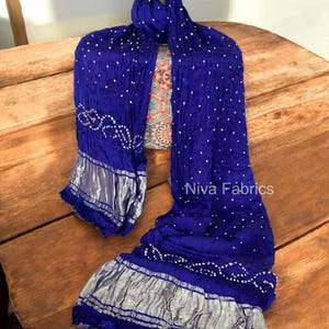 Op de afbeelding: Een koningsblauwe sjaal met witte stippen en zilveren rand. De sjaal is gedrapeerd over een houten oppervlak. De tekst "Niva Fabrics" is zichtbaar op de sjaal.
