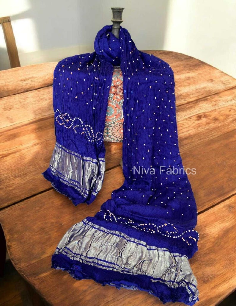 Op de afbeelding: Een koningsblauwe sjaal met witte stippen en zilveren rand. De sjaal is gedrapeerd over een houten oppervlak. De tekst "Niva Fabrics" is zichtbaar op de sjaal.