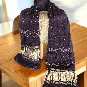 Op de afbeelding: Een donkerpaarse sjaal met witte stippen en gouden accenten is over een mannequin gedrapeerd. De sjaal heeft een textuur met decoratieve gouden rand en de tekst "Niva Fabrics".
