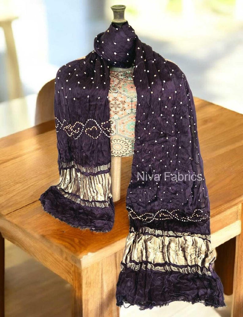 Op de afbeelding: Een donkerpaarse sjaal met witte stippen en gouden accenten is over een mannequin gedrapeerd. De sjaal heeft een textuur met decoratieve gouden rand en de tekst "Niva Fabrics".