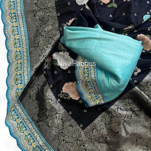 May include: A black fabric with floral patterns in white, pink, and blue. The fabric has a silver border with intricate designs and a turquoise trim. A folded turquoise fabric is also present. The text "NivaFabrics" is visible.