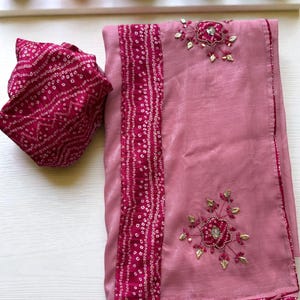 May include: A pink saree with a matching blouse piece. The saree features a dark pink border and a decorative band with a dotted pattern. Floral embroidery with gold accents adorns the saree.