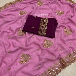 May include: A light pink saree featuring gold embroidery and sequins along the border and scattered floral motifs. A folded dark purple fabric piece with gold embroidery and the text NivaFabrics is placed on top of the pink fabric.