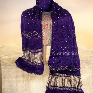 Op de afbeelding: Een dieppaarse sjaal met witte stippen en gouden randen, gedrapeerd. De sjaal wordt getoond op een paspop, waarbij de vloeiende stof en decoratieve elementen worden benadrukt. De tekst "Niva Fabrics" is zichtbaar.