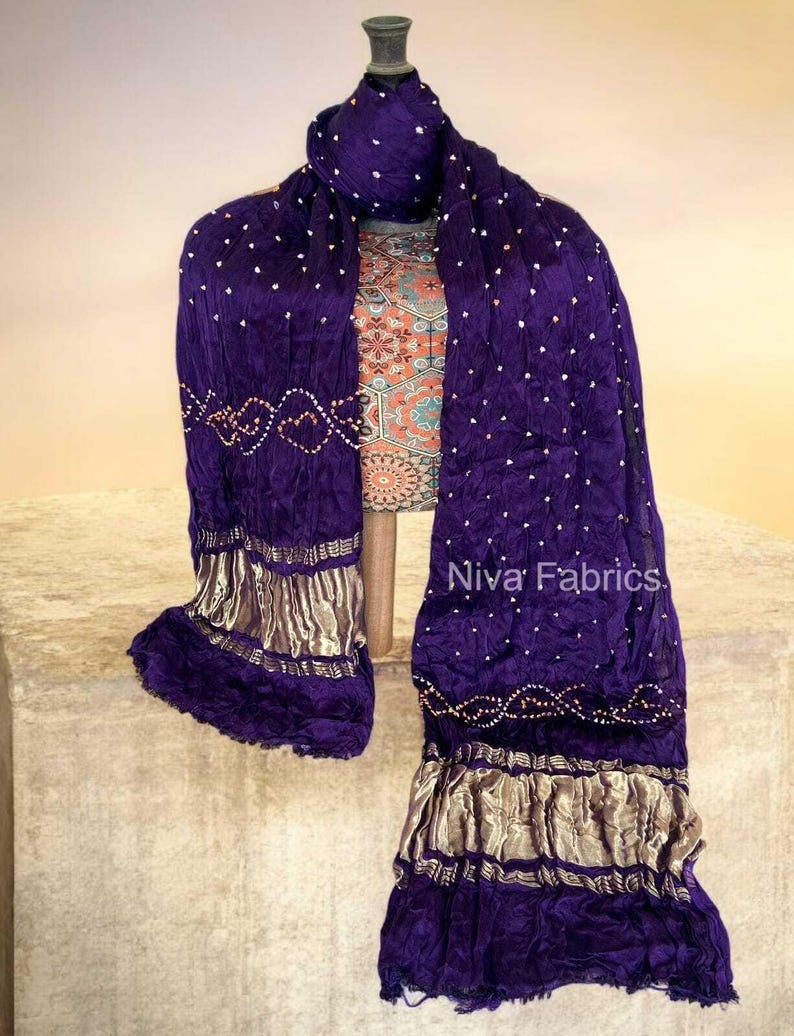 Op de afbeelding: Een dieppaarse sjaal met witte stippen en gouden randen, gedrapeerd. De sjaal wordt getoond op een paspop, waarbij de vloeiende stof en decoratieve elementen worden benadrukt. De tekst "Niva Fabrics" is zichtbaar.