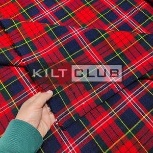 May include: Close-up of a red, navy, green, and yellow plaid fabric. The fabric has a tartan pattern with intersecting lines. The text "KILT CLUB" is visible on the fabric. A hand wearing a green sleeve is holding the fabric.