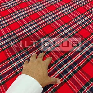 Può includere: Tessuto a quadri rossi con strisce bianche, blu navy e verdi. Il tessuto ha un motivo diagonale e il testo "KILT CLUB" è visibile. Una mano sta toccando il tessuto.